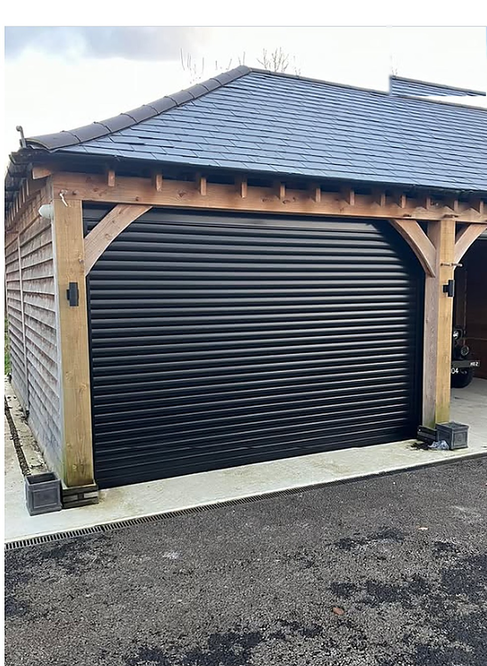 Black roller garage door fitted under an oak-framed carport — a Wilks installation
